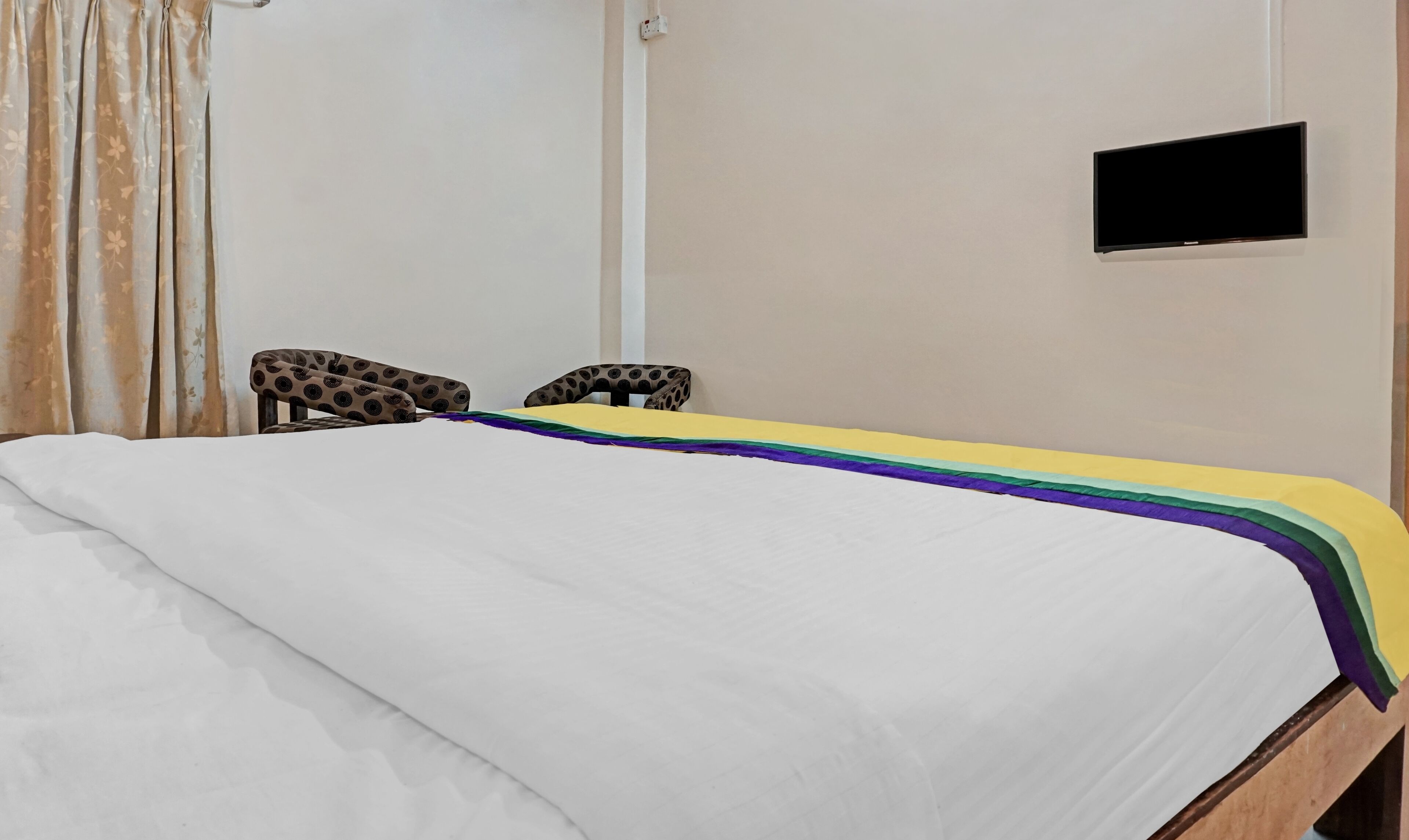 A neatly made bed with a white sheet and a colorful striped blanket. There are two chairs with a polka dot pattern. A flat-screen TV is mounted on the wall.