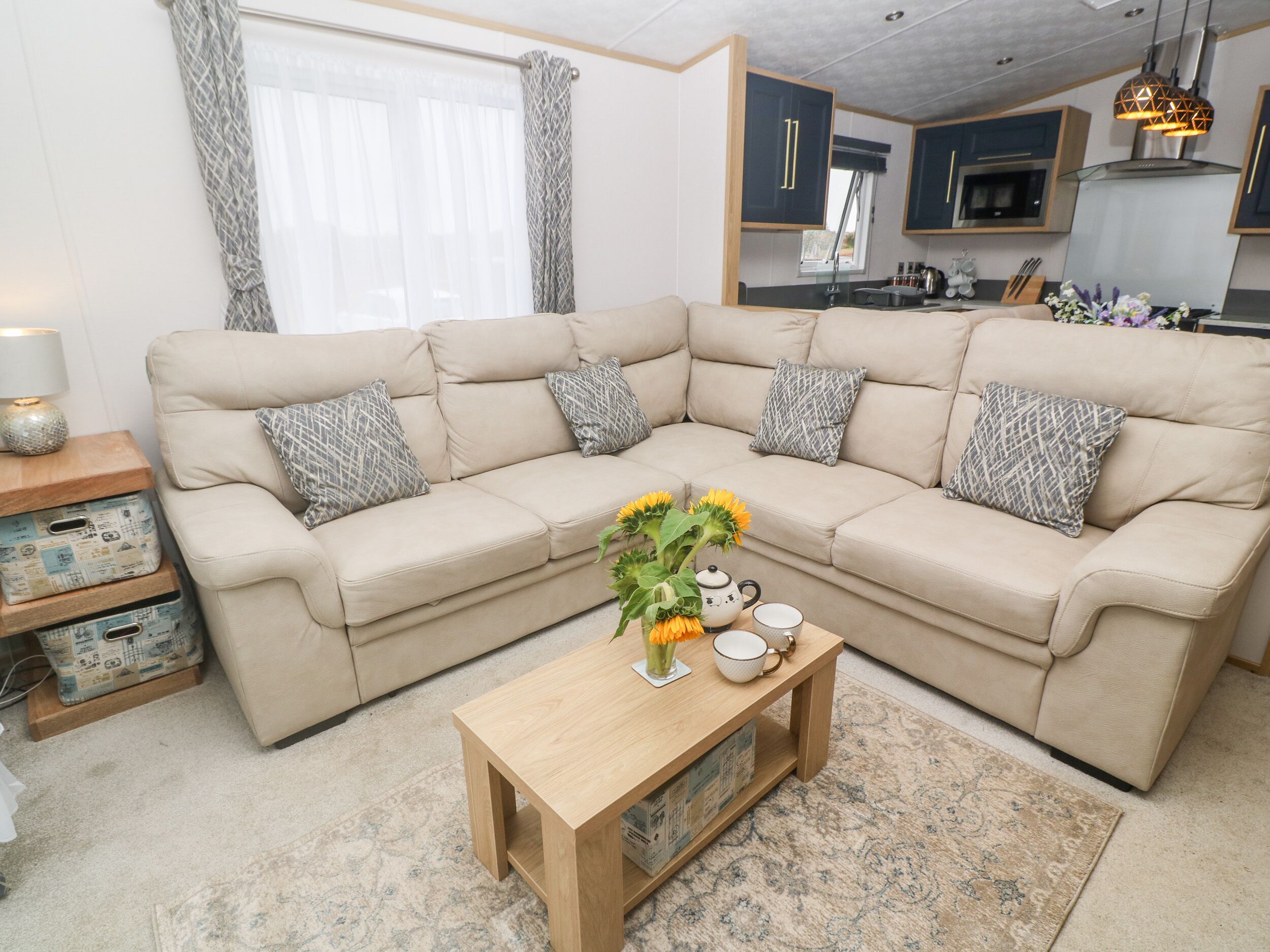 Una sala de estar con un sofá seccional color beige, cojines estampados, una mesa de centro de madera con un jarrón de girasoles y una cocina al fondo.