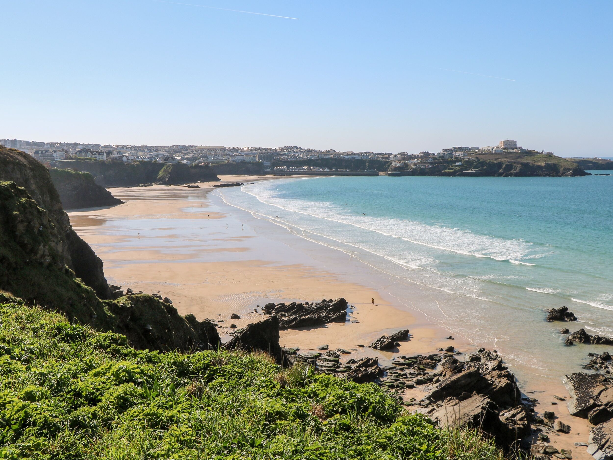 Un paysage côtier avec une plage de sable, des affleurements rocheux et une colline lointaine parsemée de bâtiments.