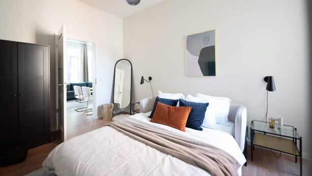 Una camera da letto con un letto, un comodino, una lampada e vista su un soggiorno.
