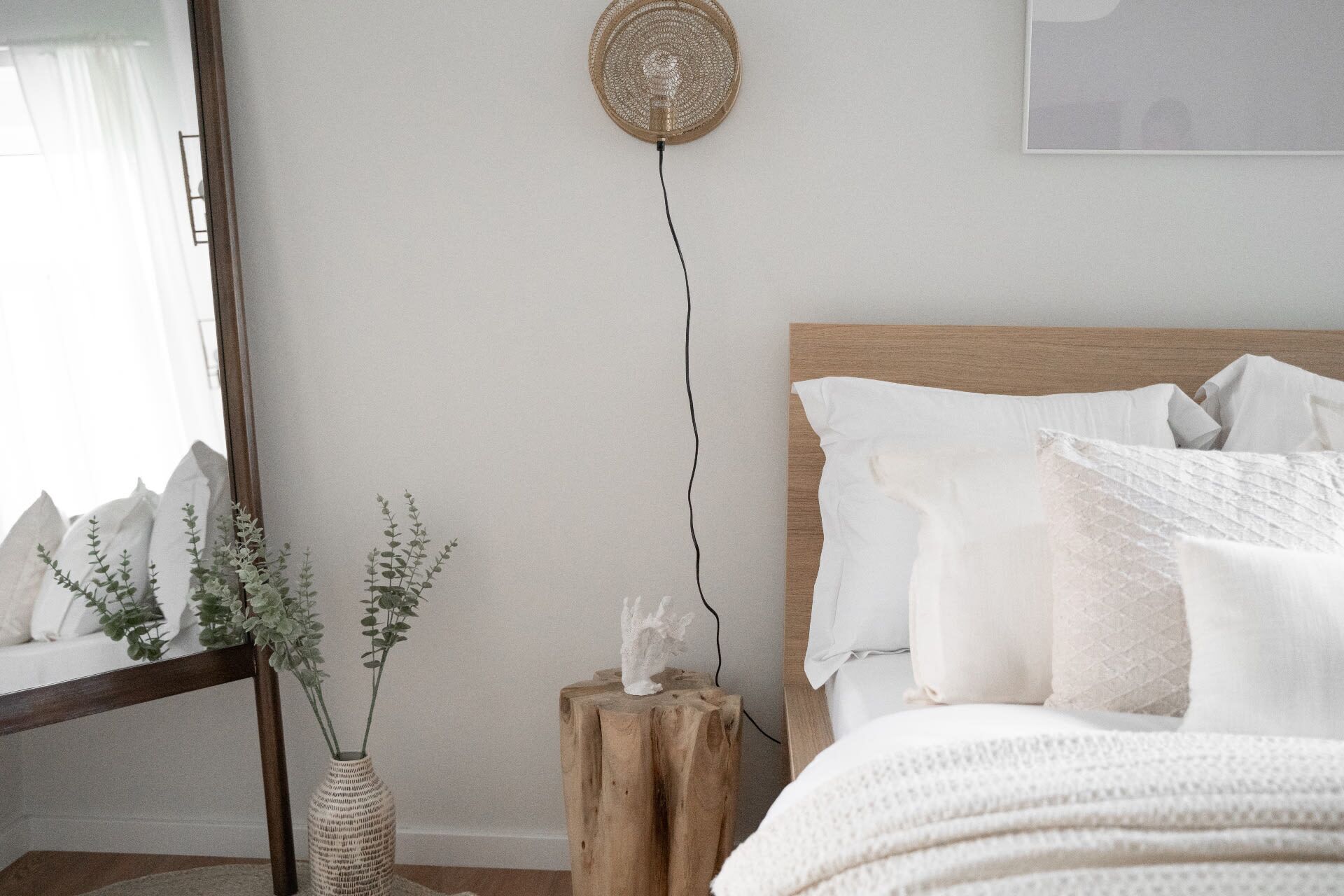 Une chambre à coucher avec une tête de lit en bois, du linge de lit blanc, une plante sur la table de chevet et un luminaire fixé au mur.