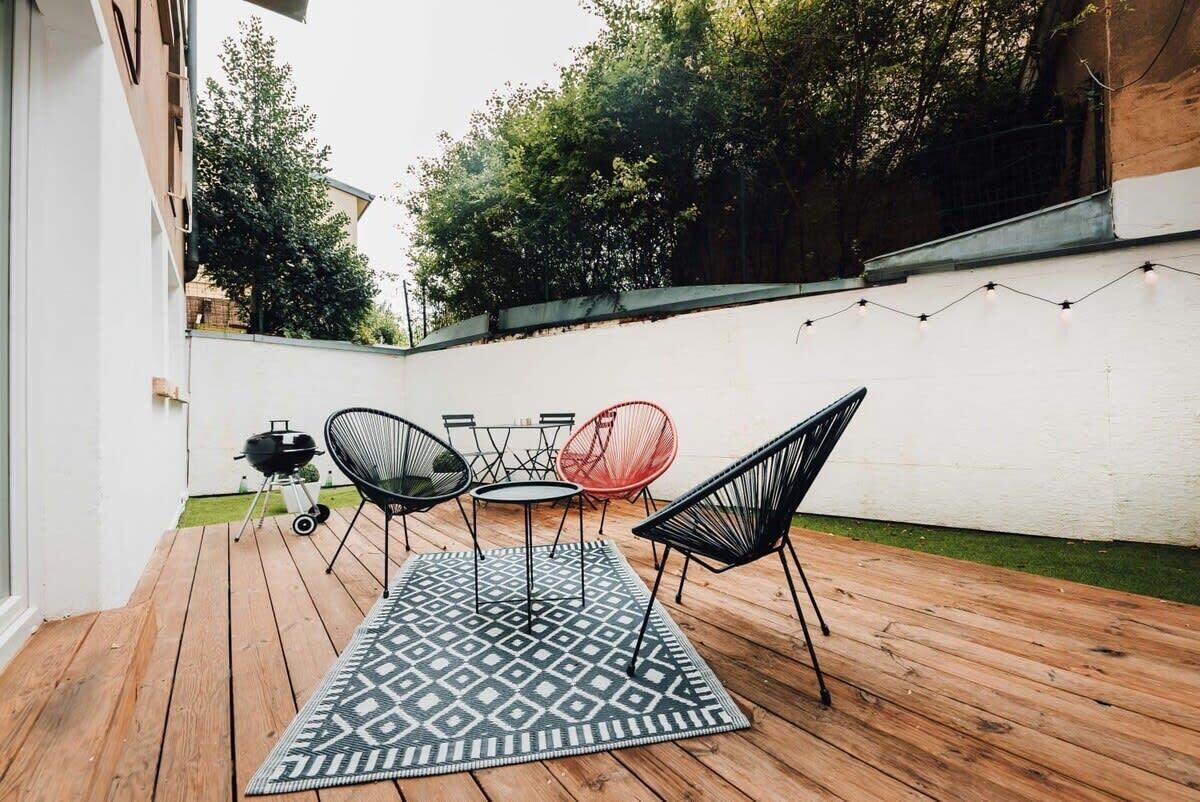 Een houten terras met een kamerkleed met patroon, zwarte en rode stoelen en een barbecue.