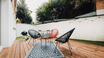 Een houten terras met een kamerkleed met patroon, zwarte en rode stoelen en een barbecue.