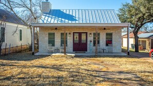 A house with a stone facade, a red door, and a covered porch with two chairs and a table. - Wine Not?- 3 BR | Hot Tub | Great for Family (Fredericksburg)
