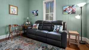 Quarto com sofá de couro preto, uma cadeira de madeira, tapete estampado e obras de arte nas paredes.