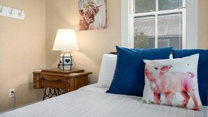 Quarto com cama, mesa de cabeceira com abajur, janela e um quadro na parede.