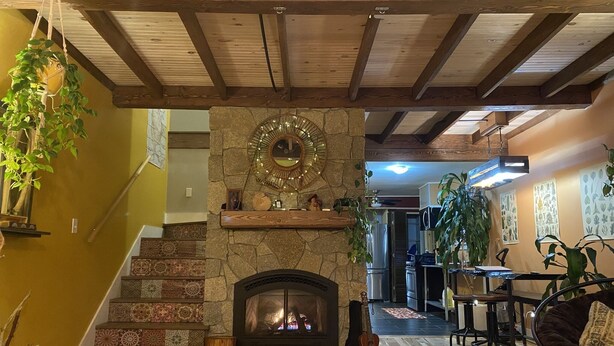 A cozy living room with a stone fireplace, wooden beams, and a staircase leading upstairs. - The Riverfront Retreat- 27 minutes to Sugarloaf! (New Portland)