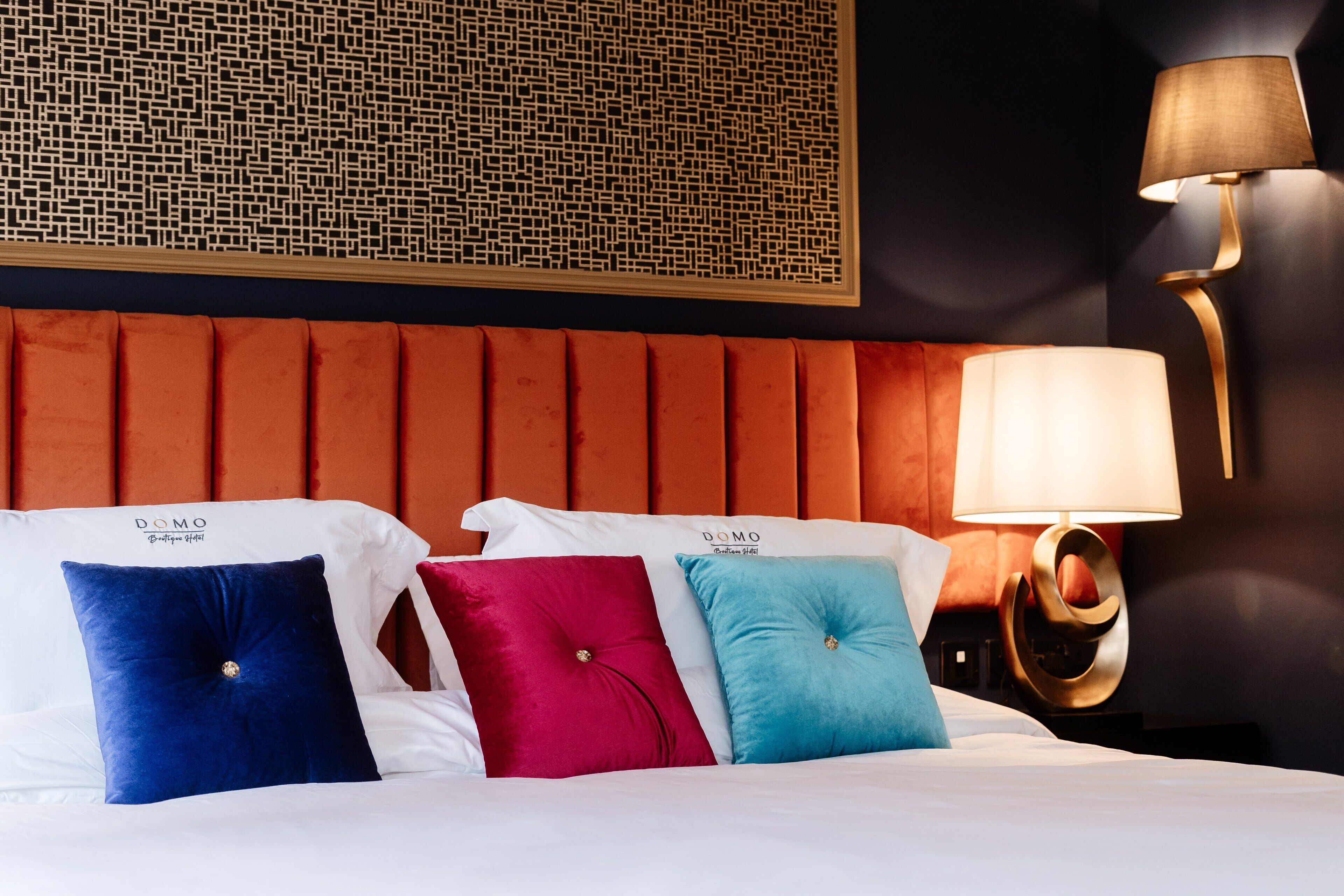Quarto de hotel com cabeceira estofada, luminária na parede e três almofadas em azul, rosa e verde-água.