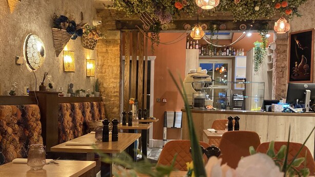 Interior de restaurante con mesas de madera, asientos acolchados, plantas colgantes e iluminación decorativa.