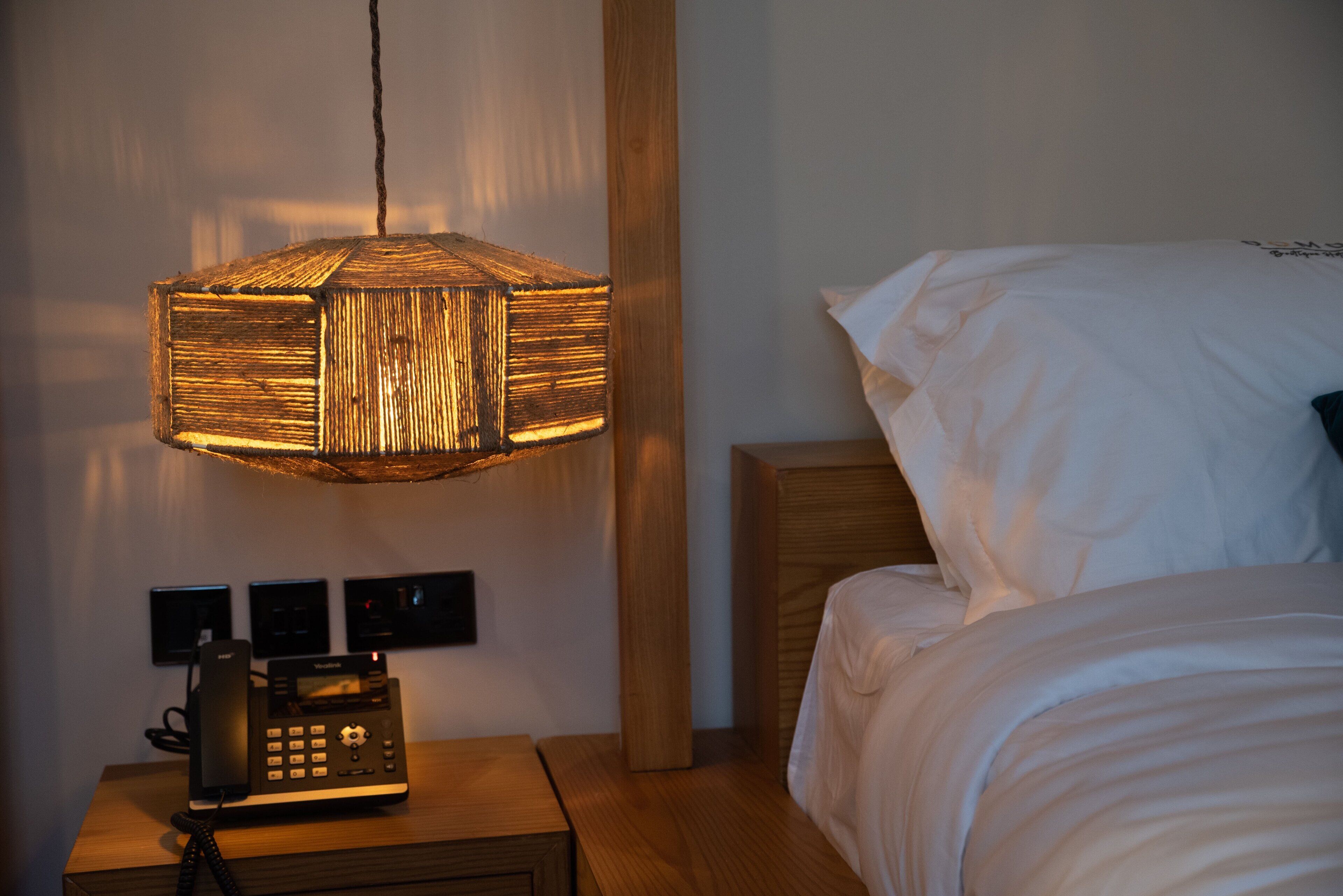 A hotel room with a wooden nightstand, a telephone, a wall-mounted light fixture, and a bed with white bedding.