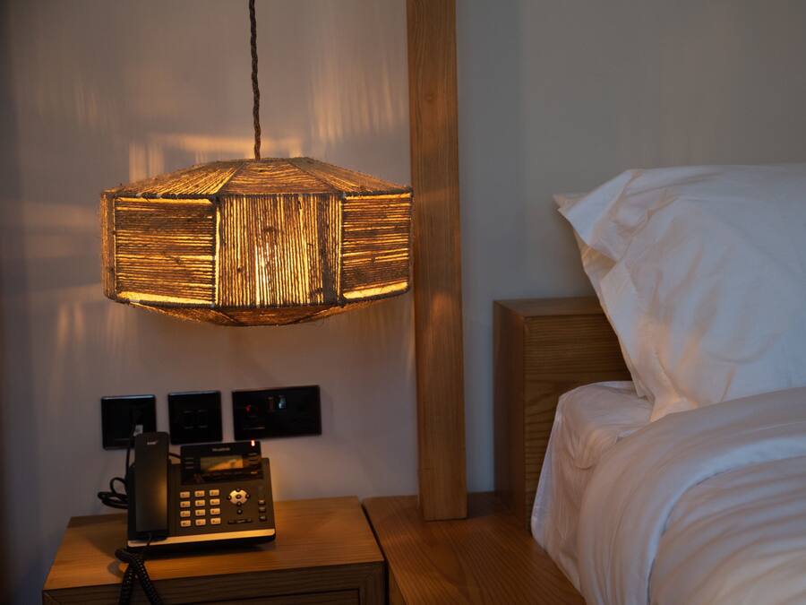 A hotel room with a wooden nightstand, a telephone, a wall-mounted light fixture, and a bed with white bedding.