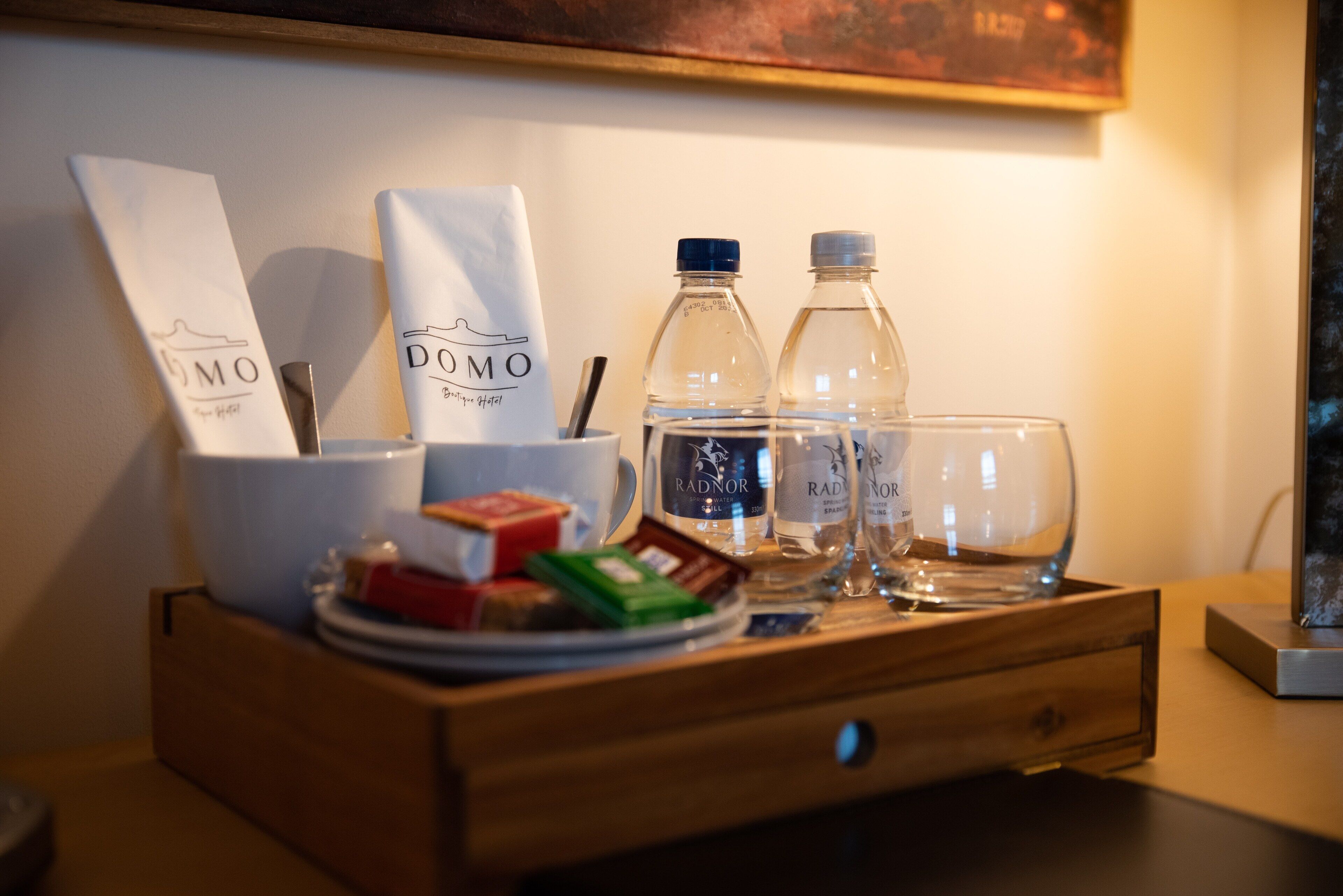 A wooden tray with a white bag labeled "DOMO", two bottles of water, a glass, and some snacks.