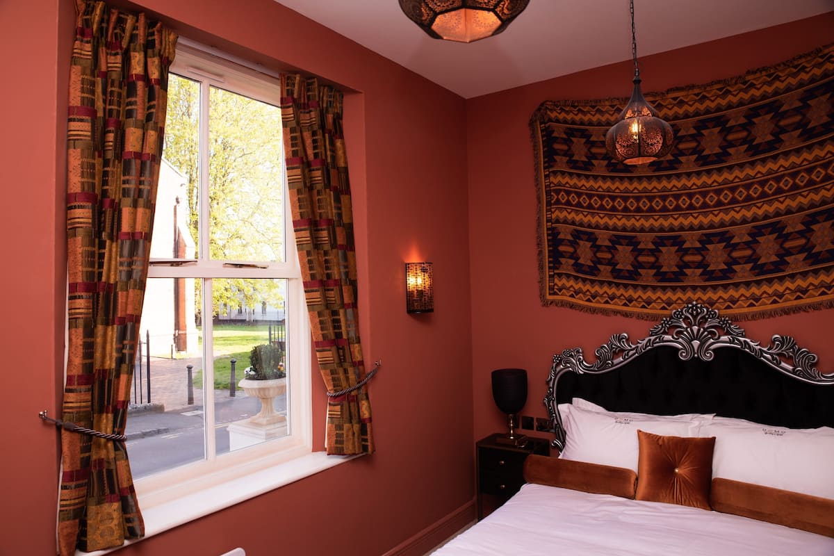 Une chambre à coucher avec un grand lit, une fenêtre avec des rideaux, une tapisserie murale et une table de chevet avec une lampe.