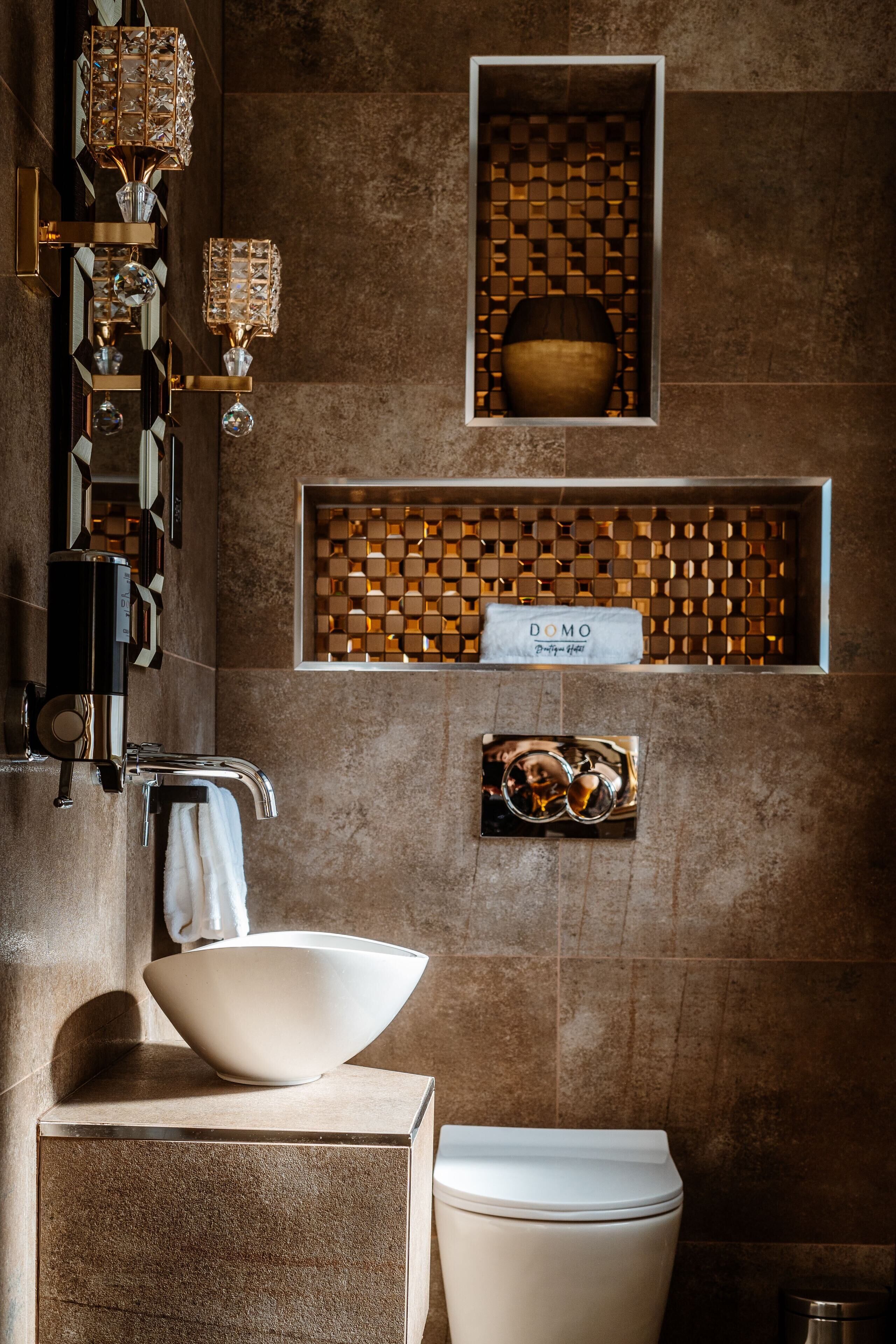 A bathroom with a large white basin, a toilet, and a wall-mounted sink. There are decorative wall sconces and a small shelf with a bowl and a sign that reads "DOMO".