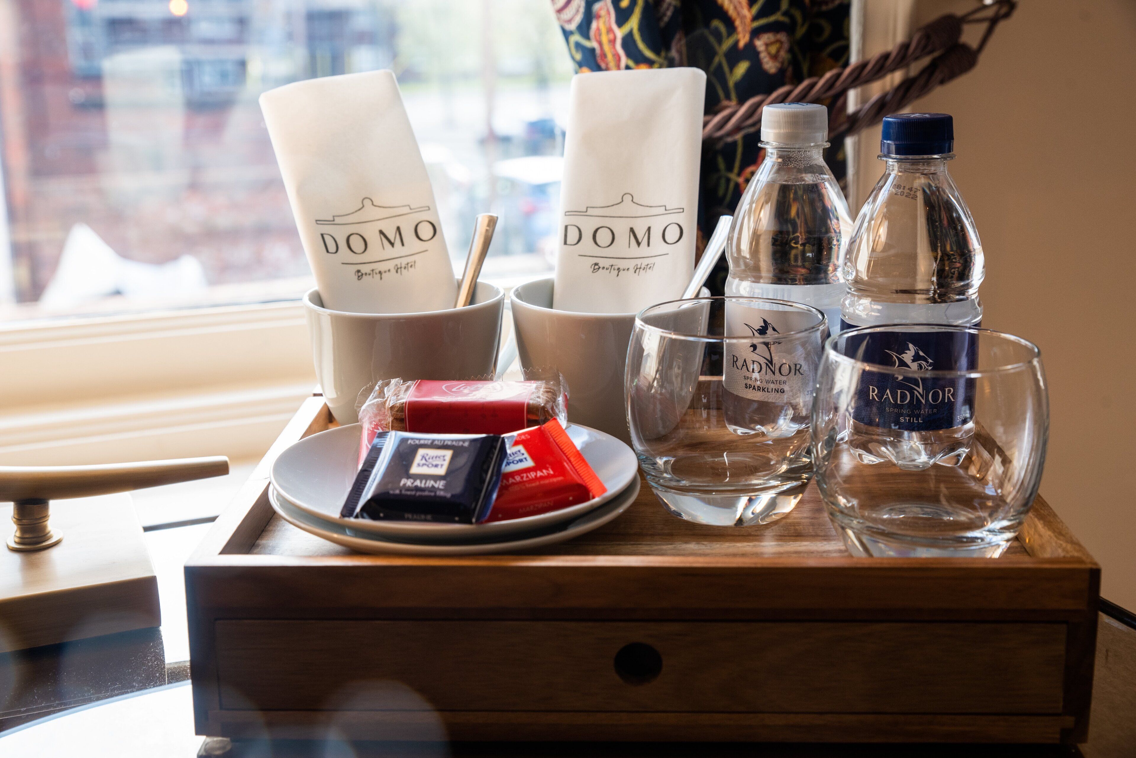 En bakke med en flaske Radnor kildevand, et glas, en tallerken med snacks og servietter med Domo-logoet.