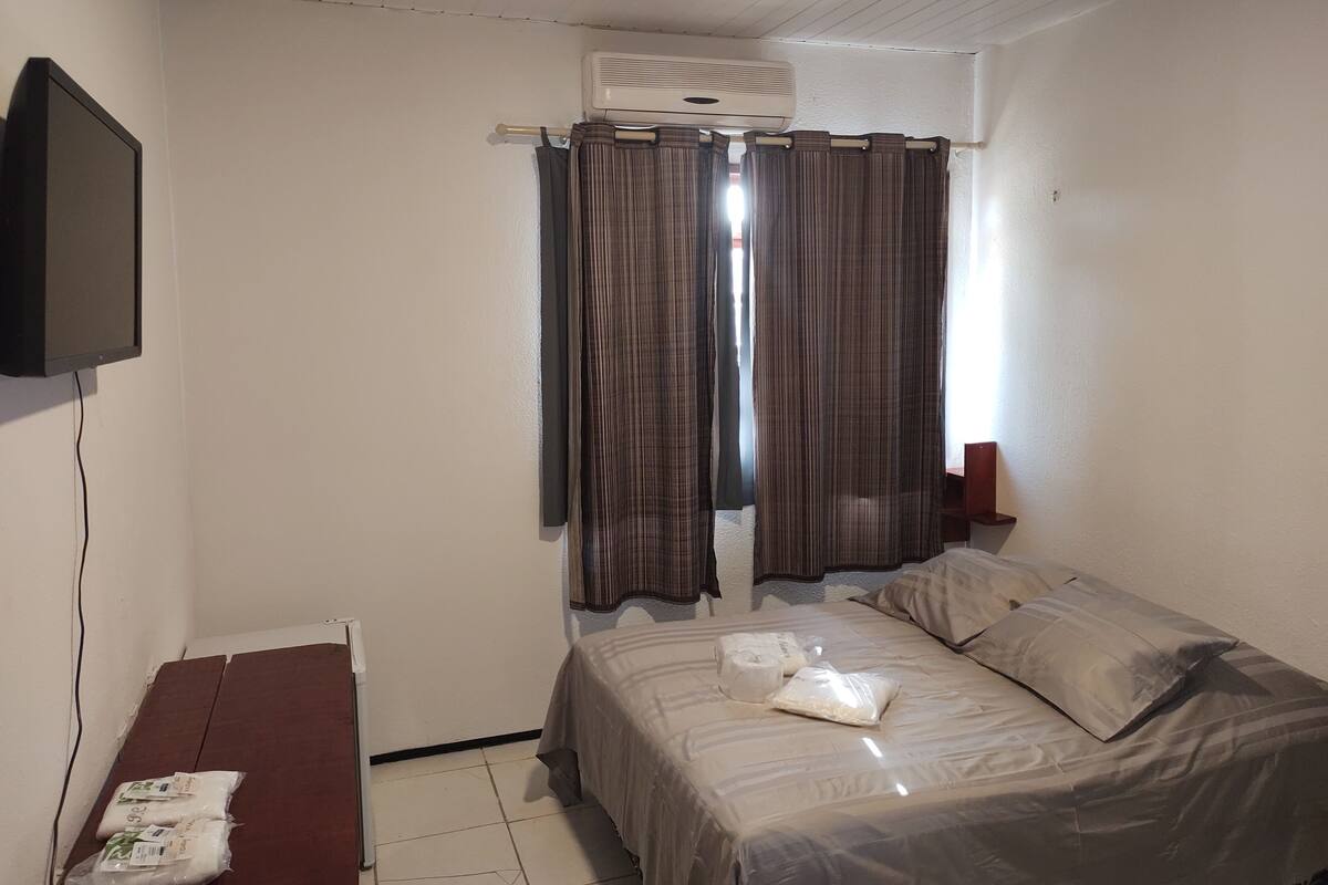 A small, sparsely furnished room with a single bed, a television mounted on the wall, and a window with curtains.