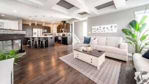A modern living room with a sofa, coffee table, and a kitchen area in the background. - Amazing Luxe Home w/ AirCon & HUGE Yard | FirePit (Edmonton)