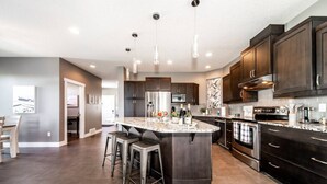 A modern kitchen with dark cabinetry, a large island with bar stools, stainless steel appliances, and pendant lighting. - Amazing Luxe Home w/ AirCon & HUGE Yard | FirePit (Edmonton)