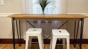 Una mesa de madera con patas de metal negro, acompañada por dos taburetes blancos con patas de metal negro.