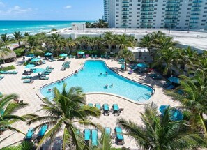 A resort pool area with multiple lounge chairs and umbrellas, surrounded by palm trees and a view of the ocean. - Lovely apartment in the beach- condo with amenities on the beach! (Hollywood)