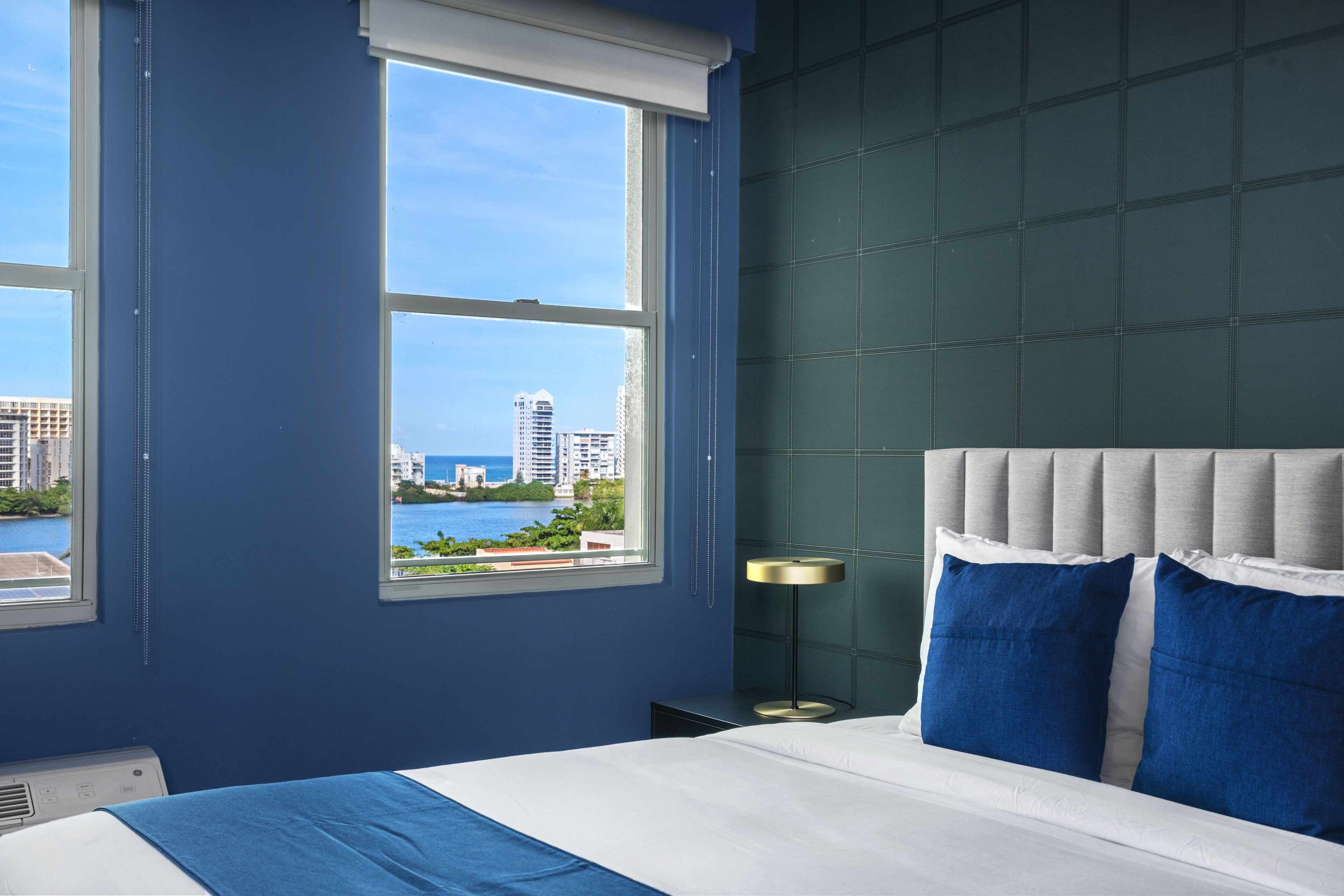 A bedroom with a large window, a bed with blue and white bedding, and a view of a cityscape and water.