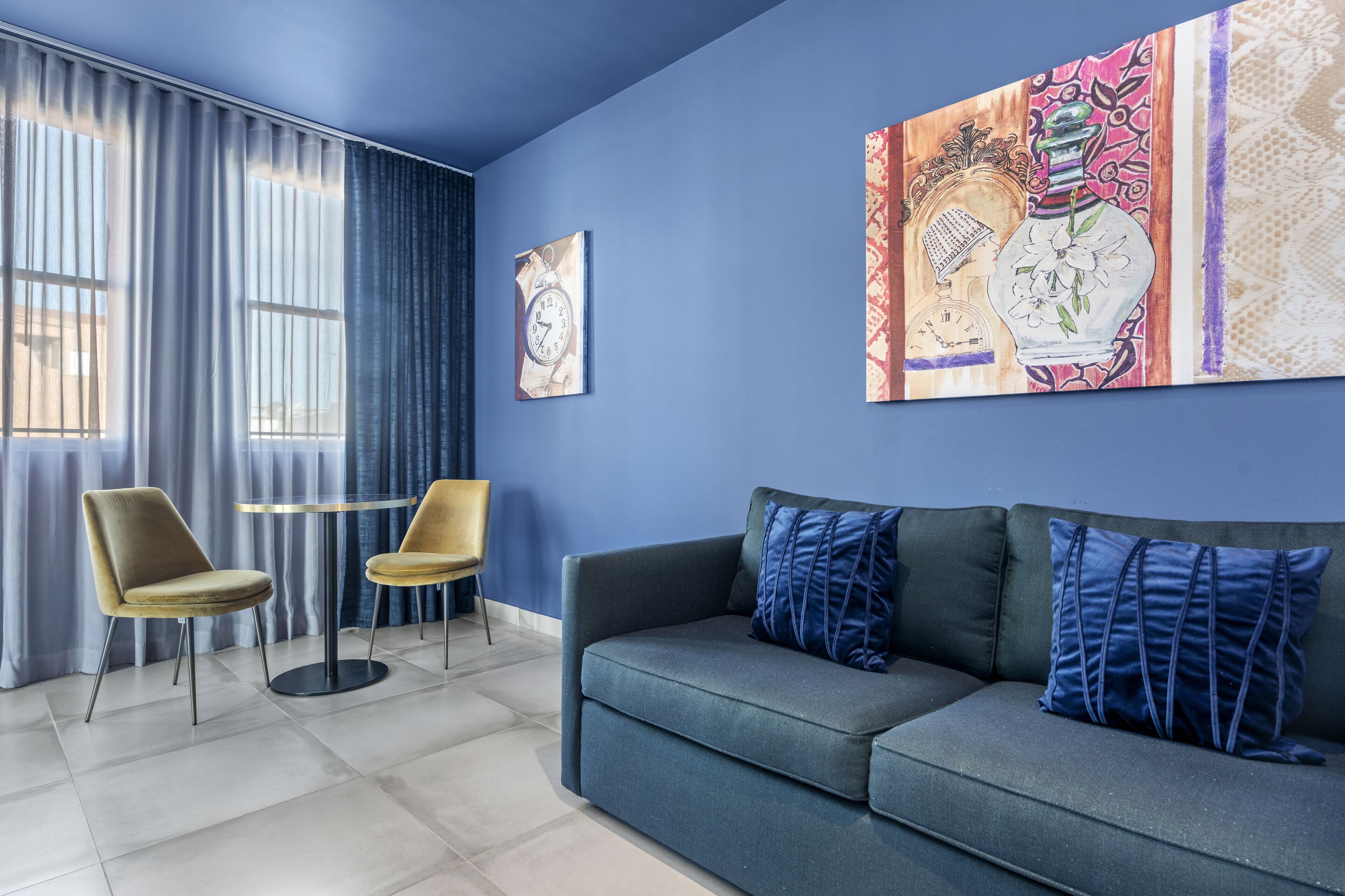 A modern living room with a blue wall, a grey sofa with blue cushions, two yellow chairs, and a round table.