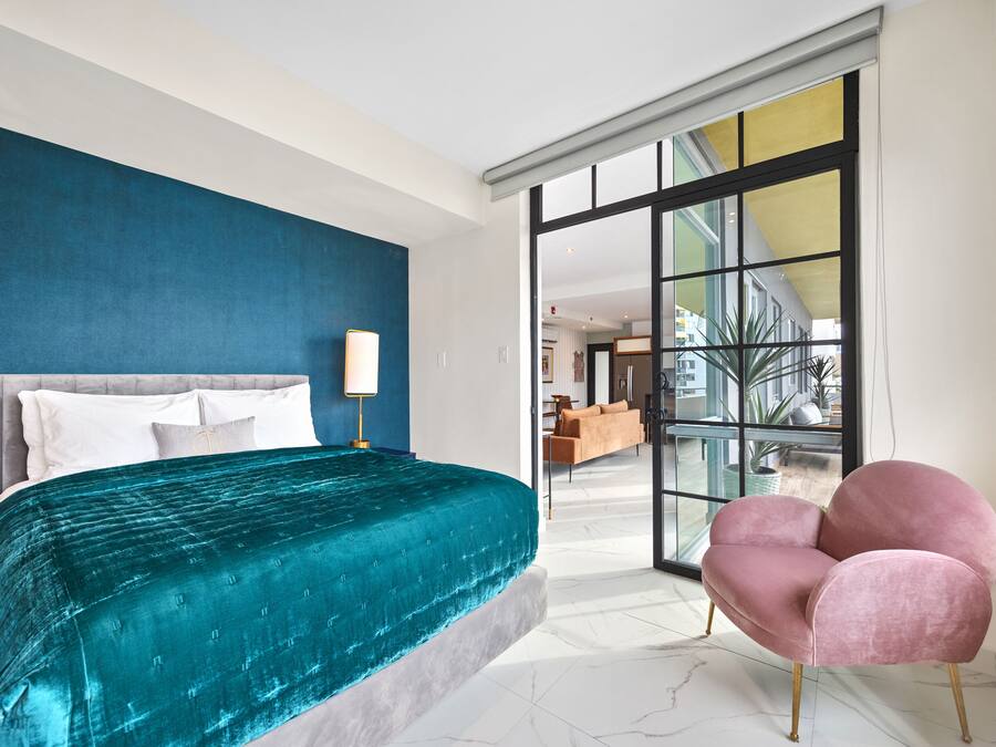 A modern bedroom with a large bed, a pink armchair, and a view through a glass door.
