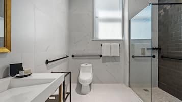 Un baño moderno con suelo de mármol blanco, cabina de ducha de vidrio, inodoro blanco y un amplio ventanal que proporciona luz natural.