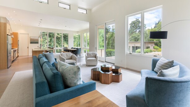 Amplia sala de estar con sofá, sillones y una mesa de centro. Hay una zona de cocina con armarios y una mesa de comedor. Ventanales grandes ofrecen luz natural y vistas al exterior.