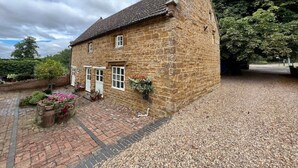 A stone house with a tiled roof, a gravel driveway, and a garden with flowers. - Braunston Manor Cottage: 4-poster beds & ensuites (Braunston)