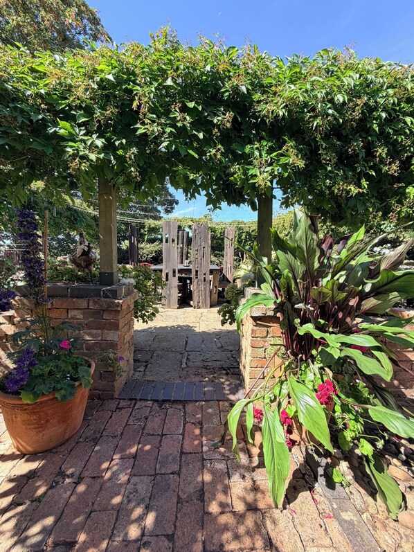 Un camí al jardí que condueix a una construcció de fusta, envoltat de vegetació abundant i plantes en testos.