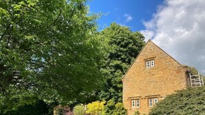 A stone house with a sloped roof, surrounded by a well-maintained lawn and various trees. - Braunston Manor Cottage: 4-poster beds & ensuites (Braunston)