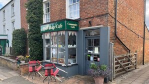 A café with a green sign, outdoor seating, and a brick building. - Braunston Manor Cottage: 4-poster beds & ensuites (Braunston)