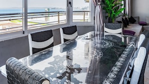 Un comedor con mesa y sillas de vidrio, ventanas amplias con vista al mar y una planta en maceta.