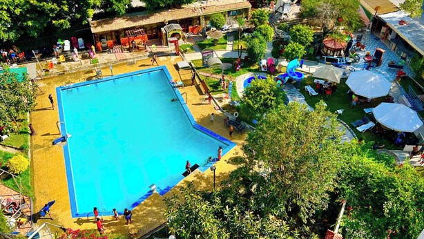 Una gran piscina al aire libre rodeada de césped, algunos árboles, pocas sombrillas y algunas personas.