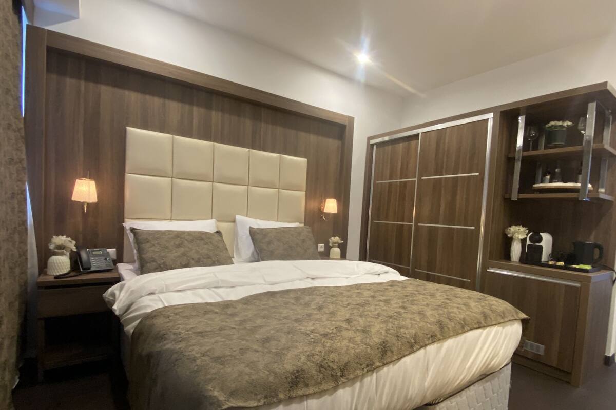 Habitación de hotel con una cama grande, una mesita de noche con lámpara, un teléfono y un pequeño jarrón con flores. Se encuentra un armario con espejo y estantes.