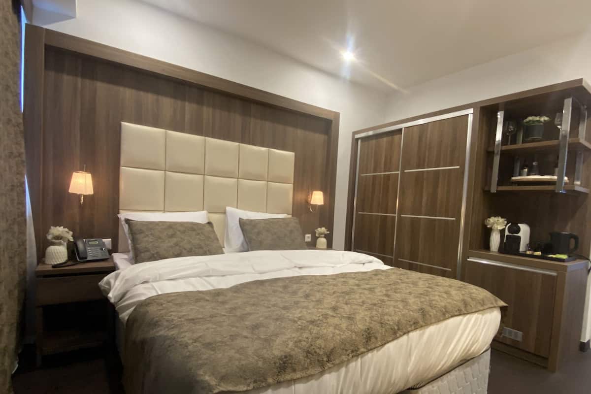 Une chambre d’hôtel avec un grand lit, une table de chevet avec une lampe, un téléphone et un petit vase avec des fleurs. Il y a aussi une armoire avec un miroir et des étagères.
