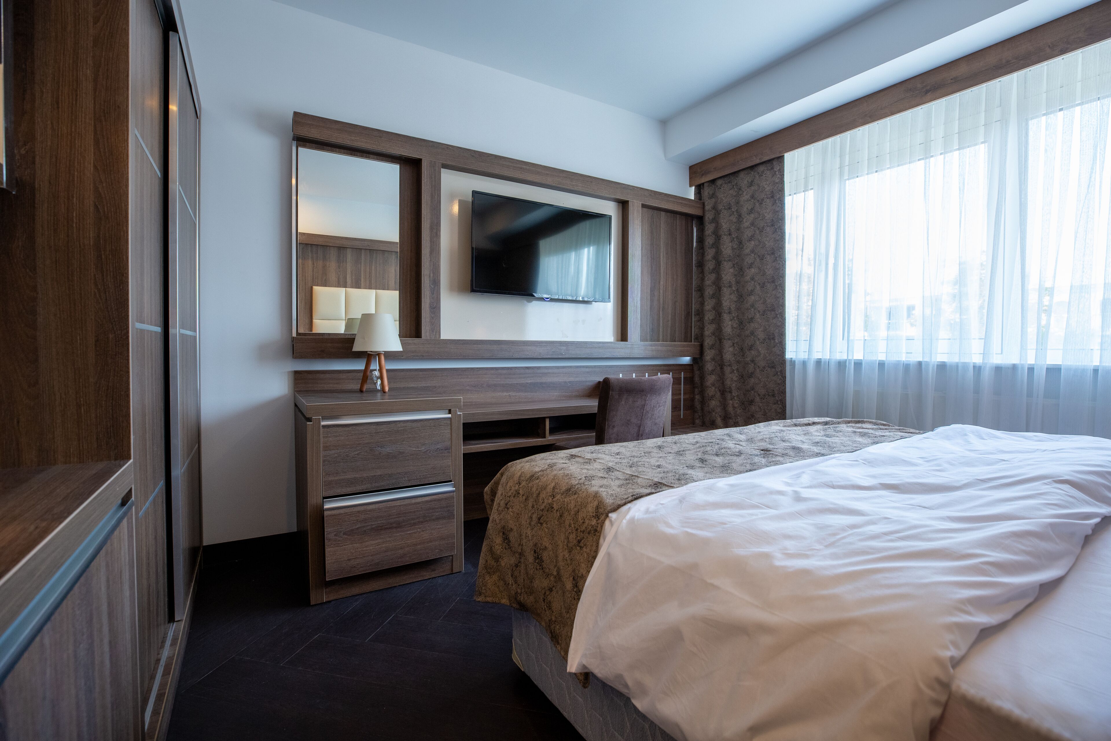 A hotel room with a bed, a desk with a chair, a TV mounted on the wall, and a window with curtains.