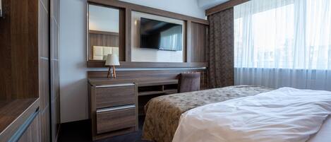 A hotel room with a bed, a desk with a chair, a TV mounted on the wall, and a window with curtains.