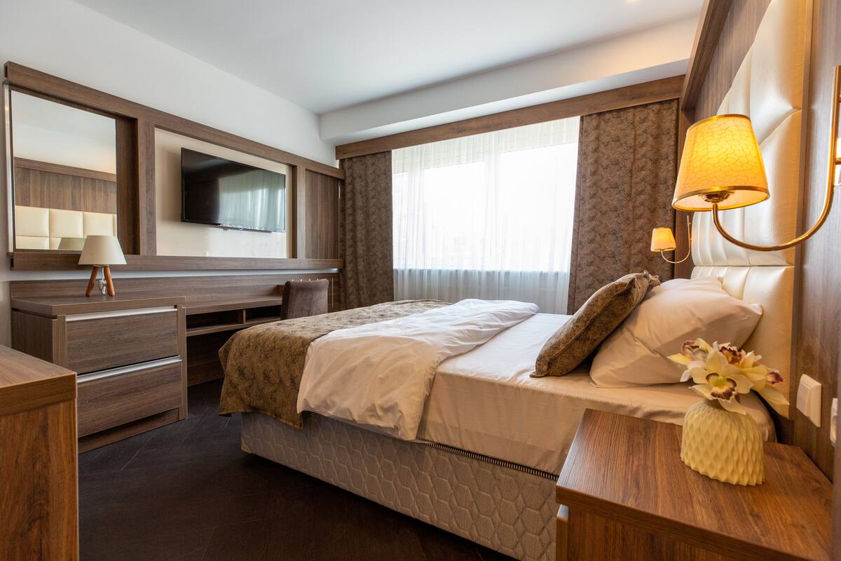 Una habitación de hotel con cama, escritorio con lámpara, televisión y un amplio ventanal con cortinas.