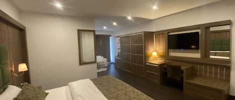 A modern hotel room with a large bed, a desk with a lamp, a TV, and a wardrobe.