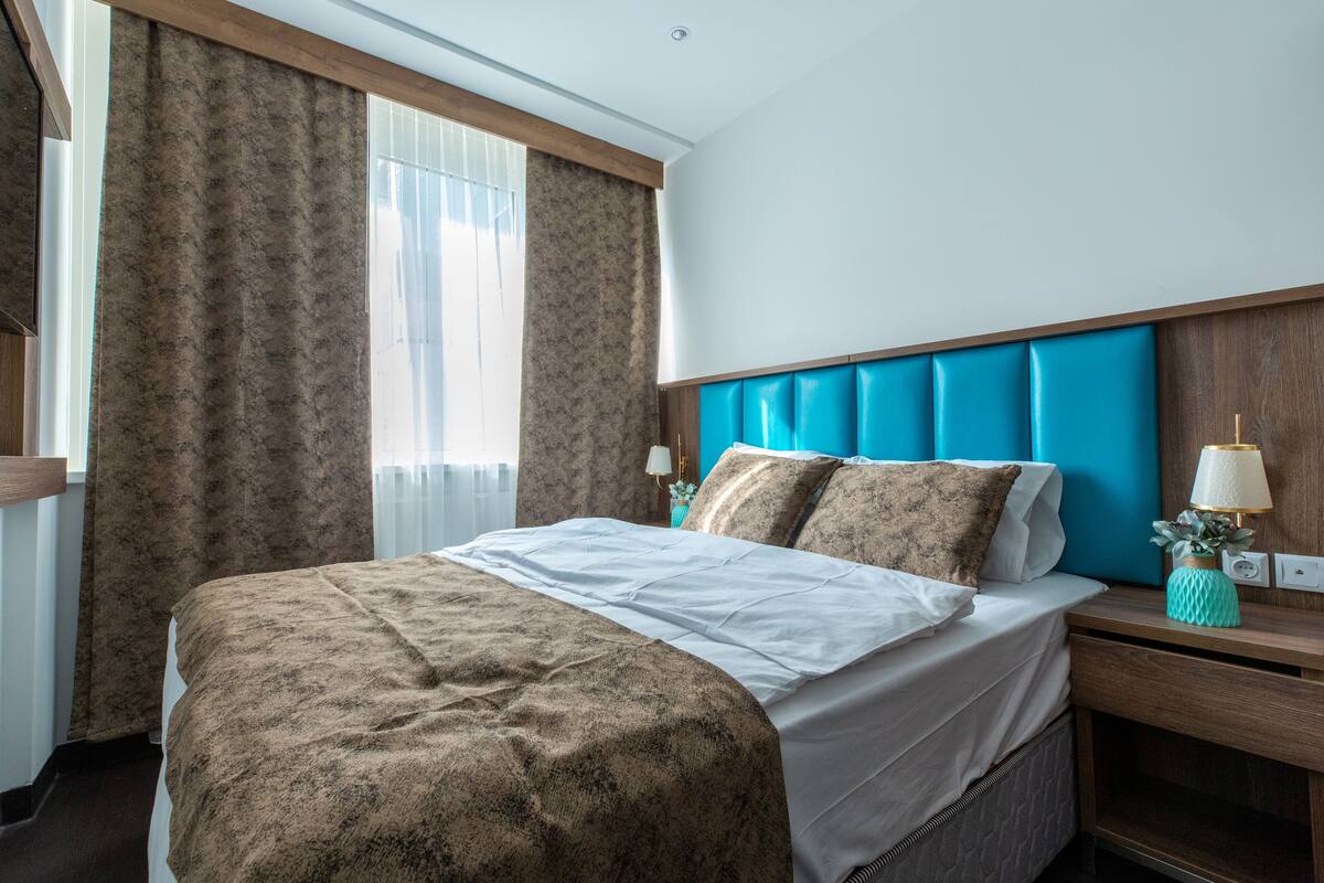 Una cama bien hecha con una colcha estampada, un cabecero con un panel azul, una mesita de noche con una lámpara y una ventana con cortinas.
