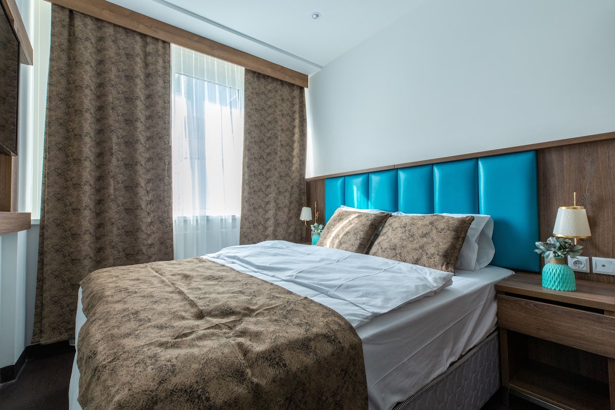 Un lit bien fait, recouvert d’une couvre-lit à motifs, une tête de lit avec un panneau bleu, une table de chevet avec une lampe et une fenêtre avec des rideaux.