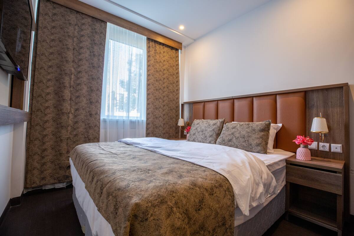 Habitación de hotel con una cama grande, un cabecero de madera, una mesita de noche con lámpara y una ventana con cortinas de tul.