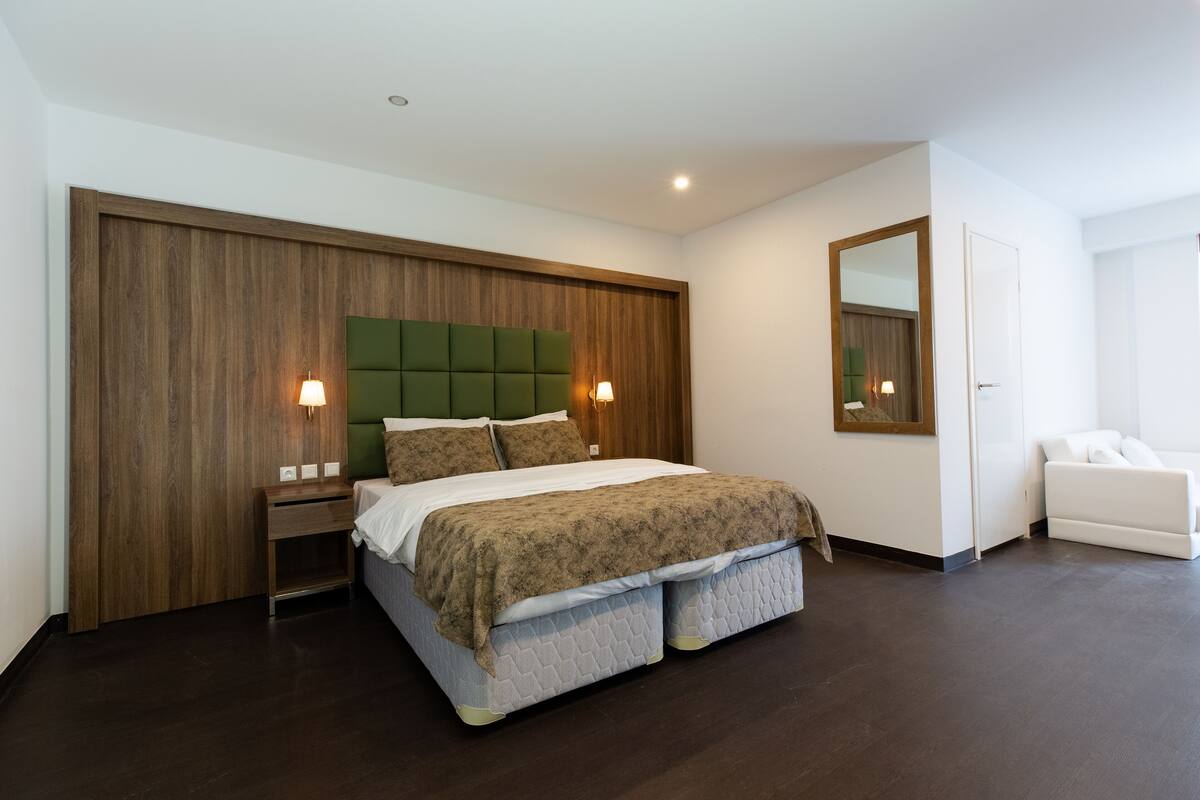 Un dormitorio moderno con una cama grande, paneles de madera, cabecera verde, lámparas de noche y una silla blanca.