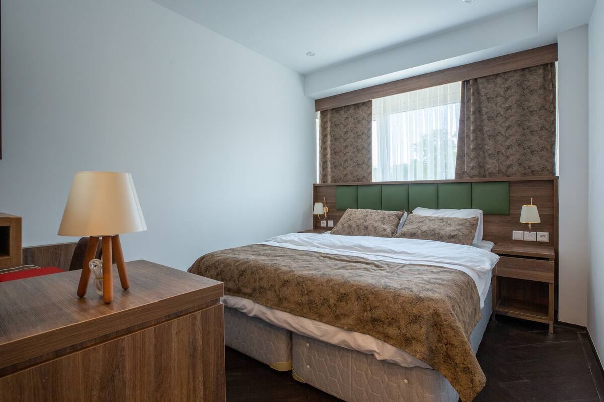 Un dormitorio con cama, lámparas de noche, una mesita de noche de madera y un escritorio con lámpara.