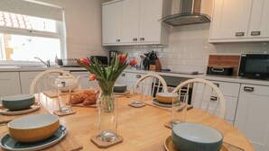 A modern kitchen with white cabinets, a round dining table set with bowls, plates, and cutlery, and a vase with flowers. - Bluebell Cottage (Bridlington)