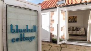 A cottage with a sign reading "bluebell cottage," open doors leading to a living area with a couch and cushions, and a tiled roof. - Bluebell Cottage (Bridlington)