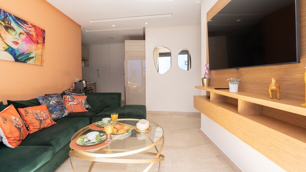 A modern living room with a green sofa, a glass coffee table with breakfast items, a large TV, and a wooden shelf with decorative items. - Majorelle - Daily rental Casablanca (Casablanca)