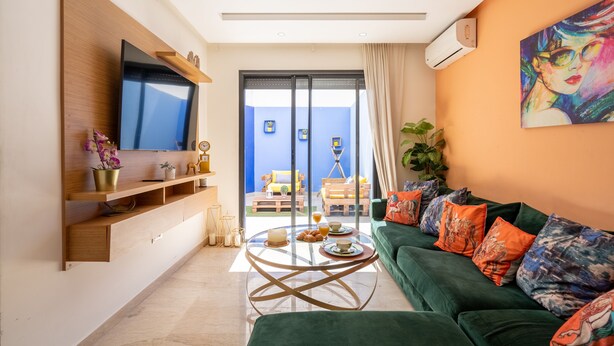 Ein modernes Wohnzimmer mit einem Flachbildfernseher, einem grünen Sofa mit bunten Kissen, einem Glastisch und Blick auf einen Balkon mit Gartenmöbeln.
